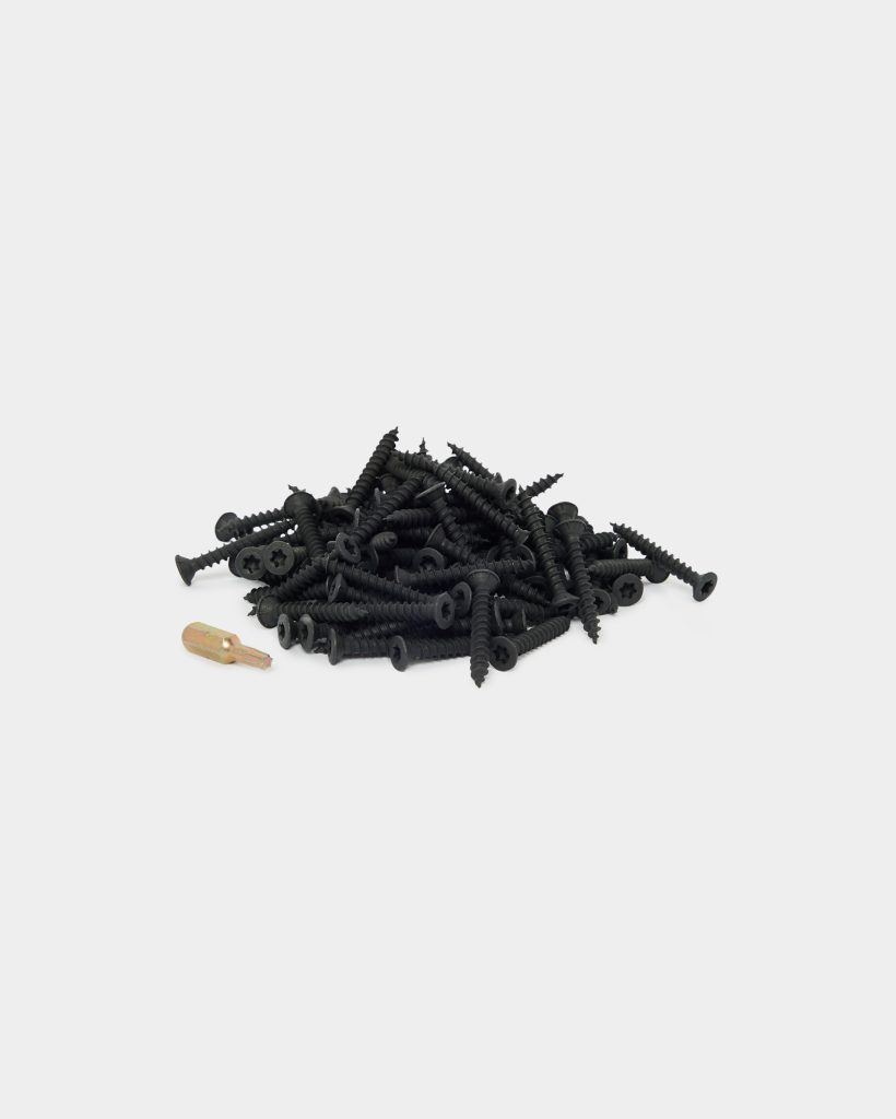 WoodUpp product image showing a small pile of black-coated wood screws with coarse threads, countersunk pan heads and star-style drives, their sharp pointed tips visible, accompanied by a single short gold-coloured screwdriver bit/adapter. Clean white background emphasizes the WoodUpp screws and matching driver.