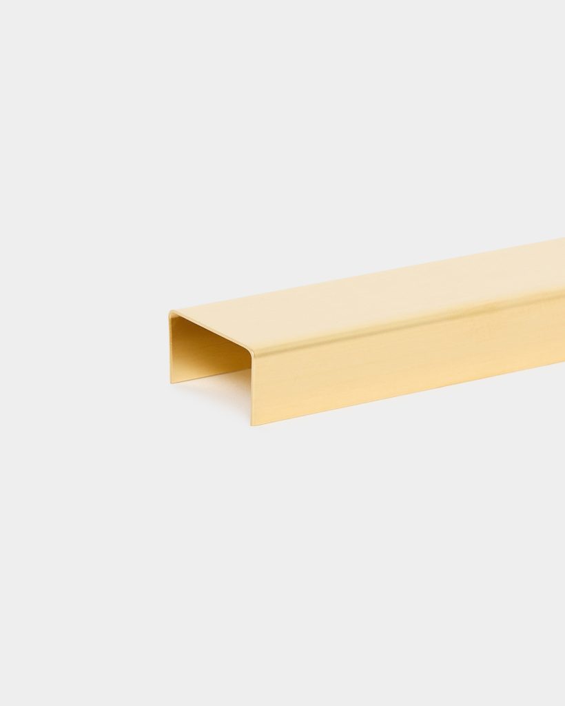 WoodUpp gold-toned U-shaped metal profile displayed on a white background. The long, hollow rectangular channel has smooth, rounded edges and a brushed metallic finish, shown diagonally across the frame — a clean, minimalist close-up of an elegant WoodUpp decorative U-profile trim piece.