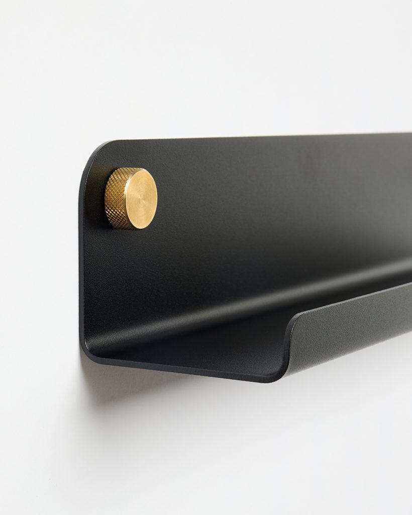 WoodUpp black wall shelf mounted on a white wall, featuring a matte black curved steel tray and a knurled brass mounting knob. Minimal floating design with rounded corners and a small front lip for holding mail, keys or small objects, a sleek storage solution from WoodUpp.