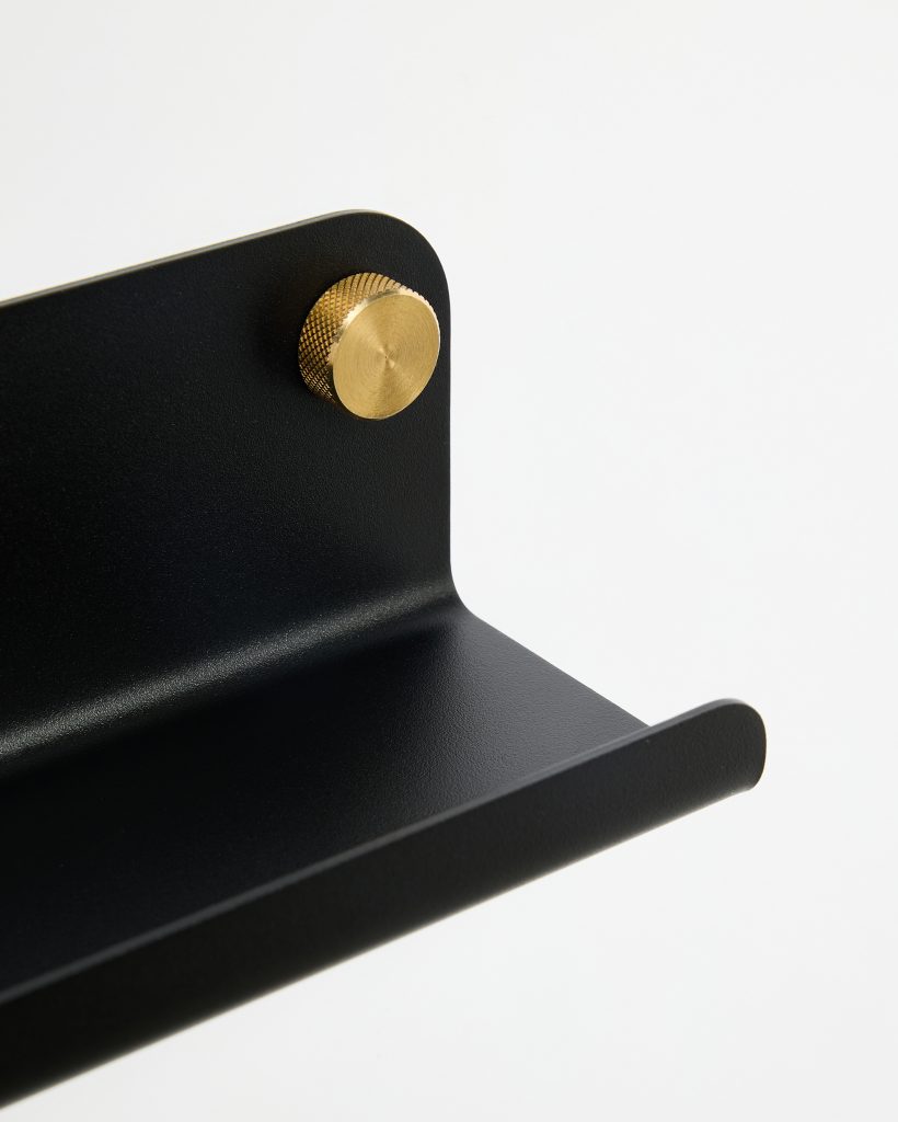 Close-up of a WoodUpp black powder-coated wall shelf showing its rounded front lip and curved back plate, secured to the wall with a single brass knurled mounting knob. The minimalist metal shelf highlights textured matte finish and decorative functional brass hardware from WoodUpp.