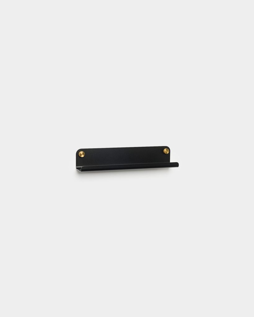 WoodUpp black wall-mounted metal shelf: a slim rectangular backplate with a rolled front lip acting as a small ledge, mounted with two exposed brass screws. Minimal, matte black finish and low-profile design suitable for holding small items like phones, keys or toiletries in modern interiors.
