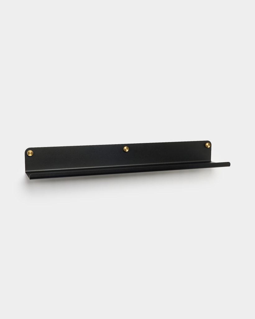 WoodUpp black metal wall shelf with a slim floating ledge and raised front lip, mounted on a white wall. The minimalist matte-black shelf features three visible brass screw heads and a subtle diagonal back plate, designed for displaying small items or framed prints.