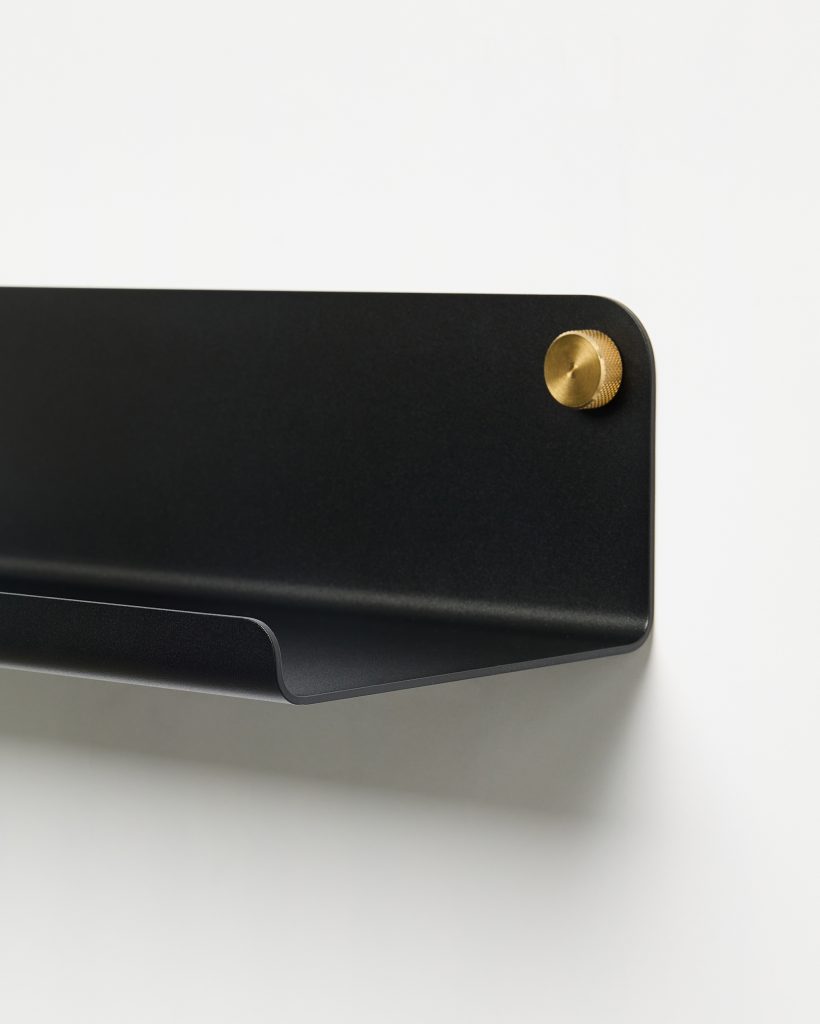Close-up of a WoodUpp black wall-mounted metal shelf with a curved front lip and matte finish, attached to a white wall by a round knurled brass mounting knob, showing its slim profile and folded-edge detail for small-item storage.