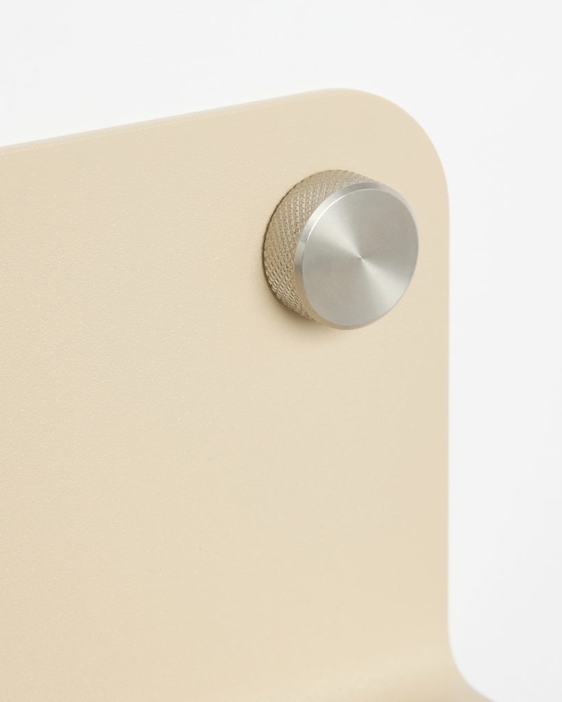 Close-up of a WoodUpp beige powder-coated metal panel with rounded corner, featuring a machined stainless-steel knurled adjustment knob. The WoodUpp product shows textured knurling and a brushed circular face on the fastener, highlighting minimalist hardware and the refined metal finish of the stand.