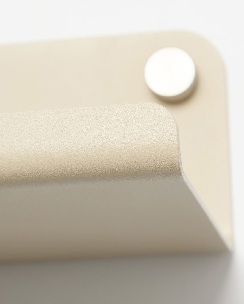 Close-up of a WoodUpp wall hook: a minimalist, powder-coated beige metal bracket with a rounded front lip and textured finish, mounted on a rectangular backplate with a polished circular fixing cap visible in the background. The image emphasizes its smooth curves, matte texture and precise metal fixing details.