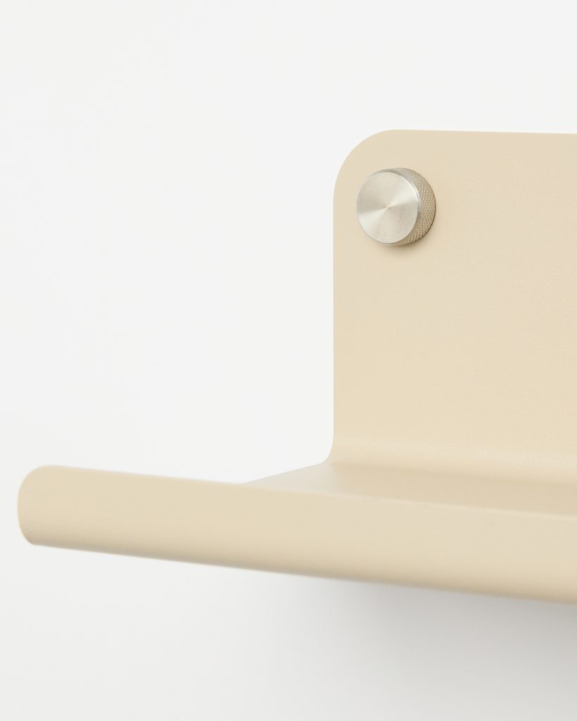 WoodUpp beige metal wall-mounted shelf bracket with a rounded front lip and smooth powder-coated finish, shown close-up attached to the wall by a brushed, knurled stainless-steel mounting knob. Minimalist, modern design highlighting the shelf's curved edge and the hardware's textured detail.