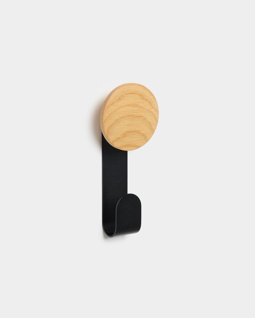 WoodUpp wall hook featuring a light oak circular peg with visible grain mounted on a slim matte-black metal strip that curves into a single downward hook. Minimal, Scandinavian-style design shown on a white wall, suitable for coats, bags, or towels.