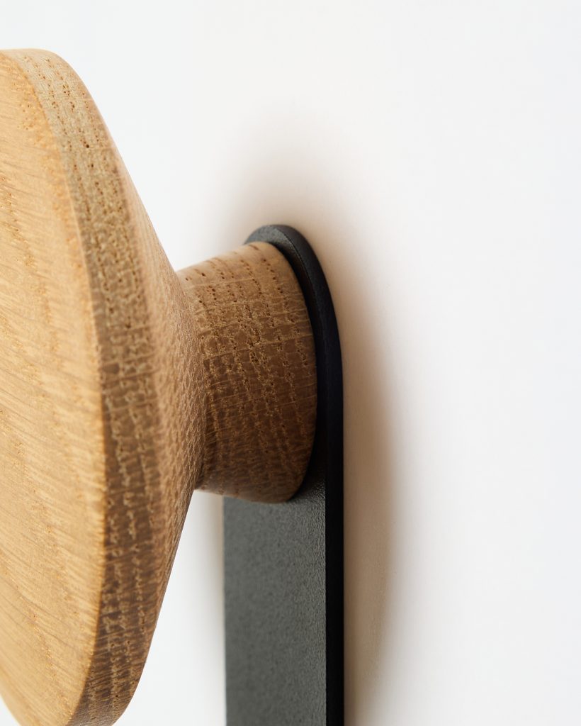 Close-up of a WoodUpp wall hook: a round oak wooden knob with visible grain and sanded texture, mounted on a slim matte-black metal bracket against a white wall, showing the knob's cylindrical base and disc-shaped front and the precise junction between wood and metal.