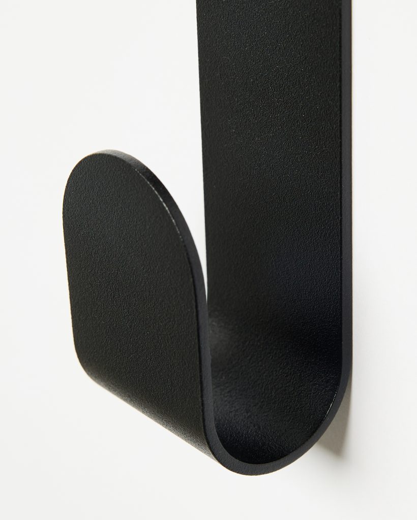 Close-up of a WoodUpp black matte wall hook mounted on a white wall. The single-piece metal hook curves into a U-shaped double-prong with rounded ends and a subtly textured finish, designed for hanging coats, towels, or bags. Minimal, modern profile in flat black.