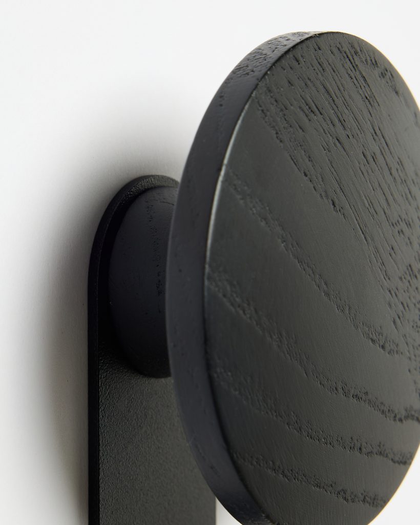 Close-up of a WoodUpp round wooden wall hook: a matte black circular ash-wood disc with visible grain, attached to a slim black metal mounting bracket and peg, shown mounted on a white wall. The product highlights textured wood grain and minimalist, modern hook design.