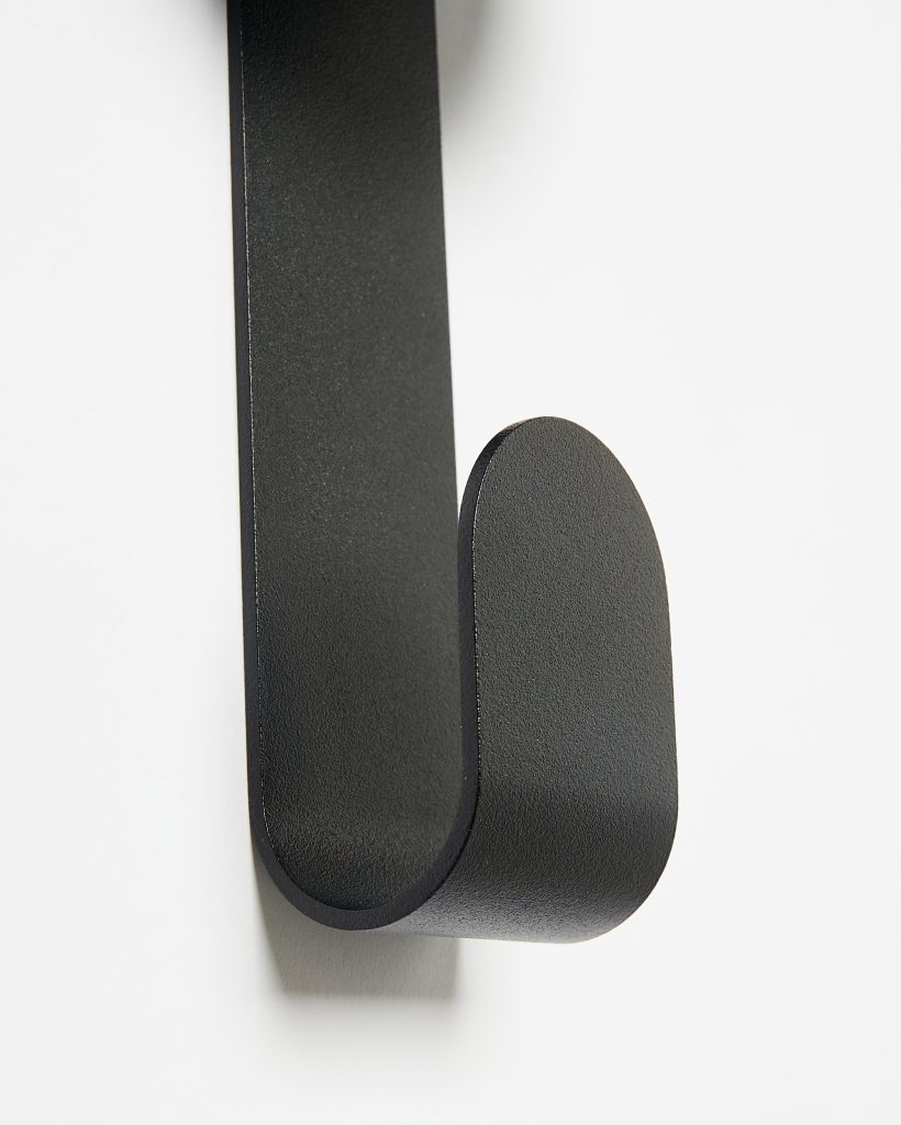 Close-up of a WoodUpp matte black wall hook: a single U-shaped, rounded metal hook with a fine textured finish and smooth curved tip, mounted on a white wall. Minimal, sturdy design for hanging coats, bags or towels in entryways and bathrooms.