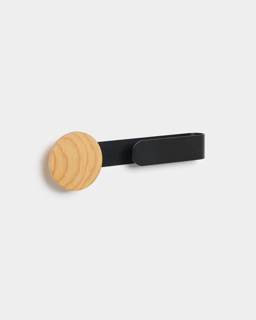 WoodUpp wall hook featuring a round natural oak disc mounted on a slim matte black metal strap that curves into a minimal hanging loop. The simple Scandinavian-style hook shows visible oak grain and a flat black powder-coated steel arm, suitable for coats, towels or bags.