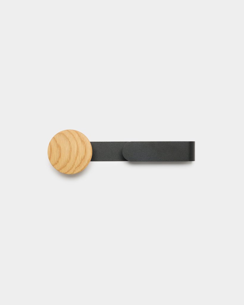 WoodUpp minimalist wall hanger featuring a round pale oak wooden knob mounted on a flat matte black leather strap loop; circular wood grain visible on left, strap extends horizontally to right with rounded end, photographed against a clean white background.