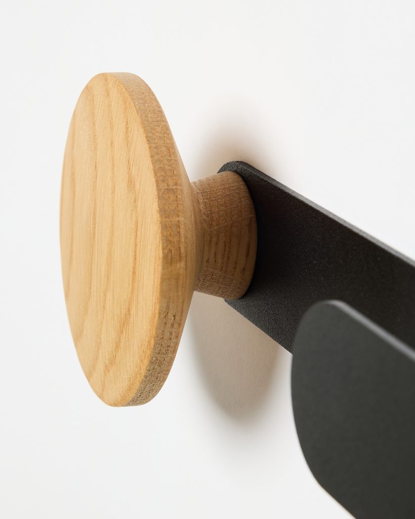 WoodUpp wall hook featuring a round oak wooden peg with visible grain and short stem, mounted on a matte black metal wall bracket; the minimalist coat hook shows a circular wooden disc head and a curved black metal arm, photographed against a white wall.