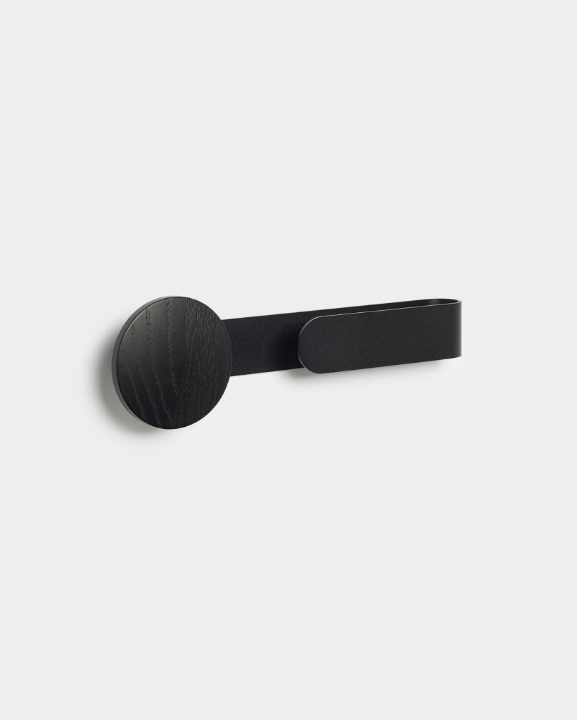 Minimalist WoodUpp wall hook featuring a matte black round wooden peg with visible grain, paired with a slim black metal strap forming a secondary horizontal hook. The compact, modern accessory mounts flush to the wall, offering concealed fixings and a sleek Nordic-inspired aesthetic.