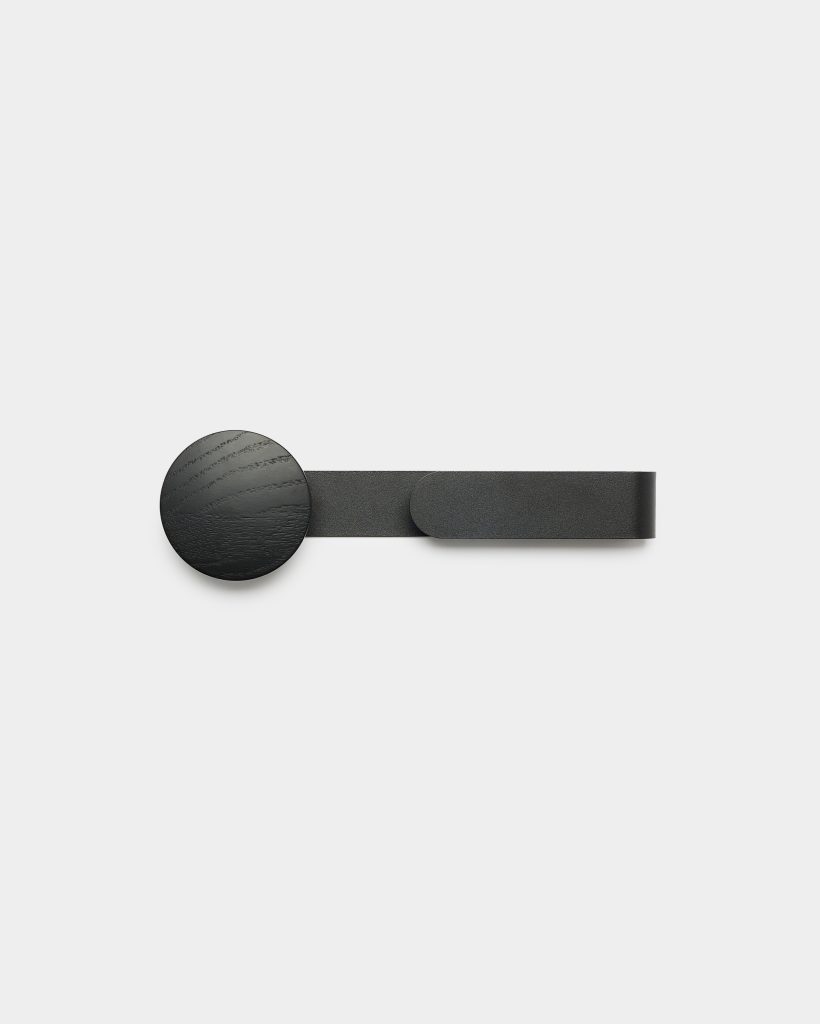 Minimalist WoodUpp wall hook: a matte black round wooden knob with visible grain mounted on a slim horizontal black metal strap that loops at the right, photographed against a clean white background, showcasing simple Scandinavian design and a textured finish, ideal for coats or accessories.