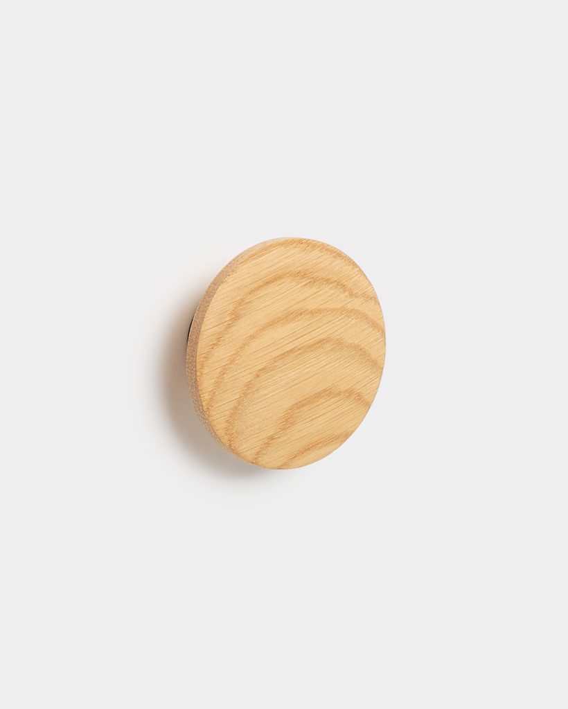 WoodUpp round wooden wall hook: a single circular oak peg with visible wood grain and smooth, light finish, mounted on a white wall. The minimalist wooden knob casts a soft shadow and exemplifies WoodUpp's simple, natural design for hanging coats, towels, or accessories.