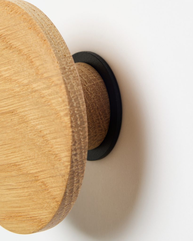 WoodUpp wooden wall hook: close-up of a round oak peg with visible grain and a thick circular face, mounted on a slim cylindrical stem and fixed to the white wall with a black metal mounting plate. Soft shadow emphasizes texture and minimalist design.