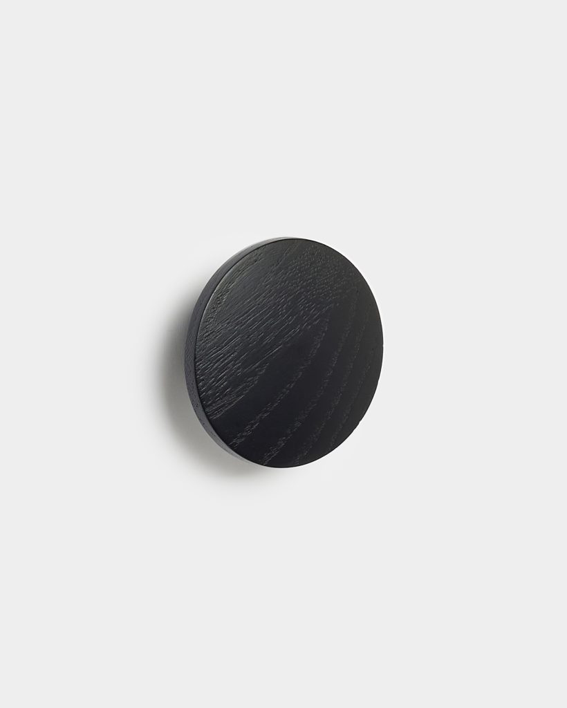 WoodUpp round wall hook: a single circular black wooden peg with visible wood grain and slim profile, mounted on a white wall. The minimalist WoodUpp coat hook shows subtle texture, smooth edges and a soft shadow, highlighting its matte finish and compact design.