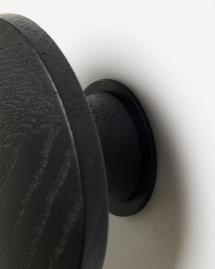 WoodUpp black wooden wall hook shown in close-up: a round, slightly beveled disk with visible wood grain and matte black finish, attached by a short cylindrical stem and flat circular base mounting plate, fastened to a white wall—simple, modern coat hook/peg design.