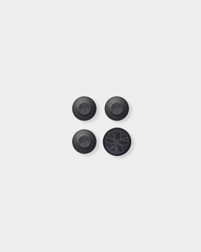 WoodUpp black thumbstick caps photographed on white background: four round silicone controller grips arranged in a square — three dome-shaped caps with an embossed radial WoodUpp logo, plus one larger flat, textured cap featuring a detailed tree‑ring pattern for added durable grip.