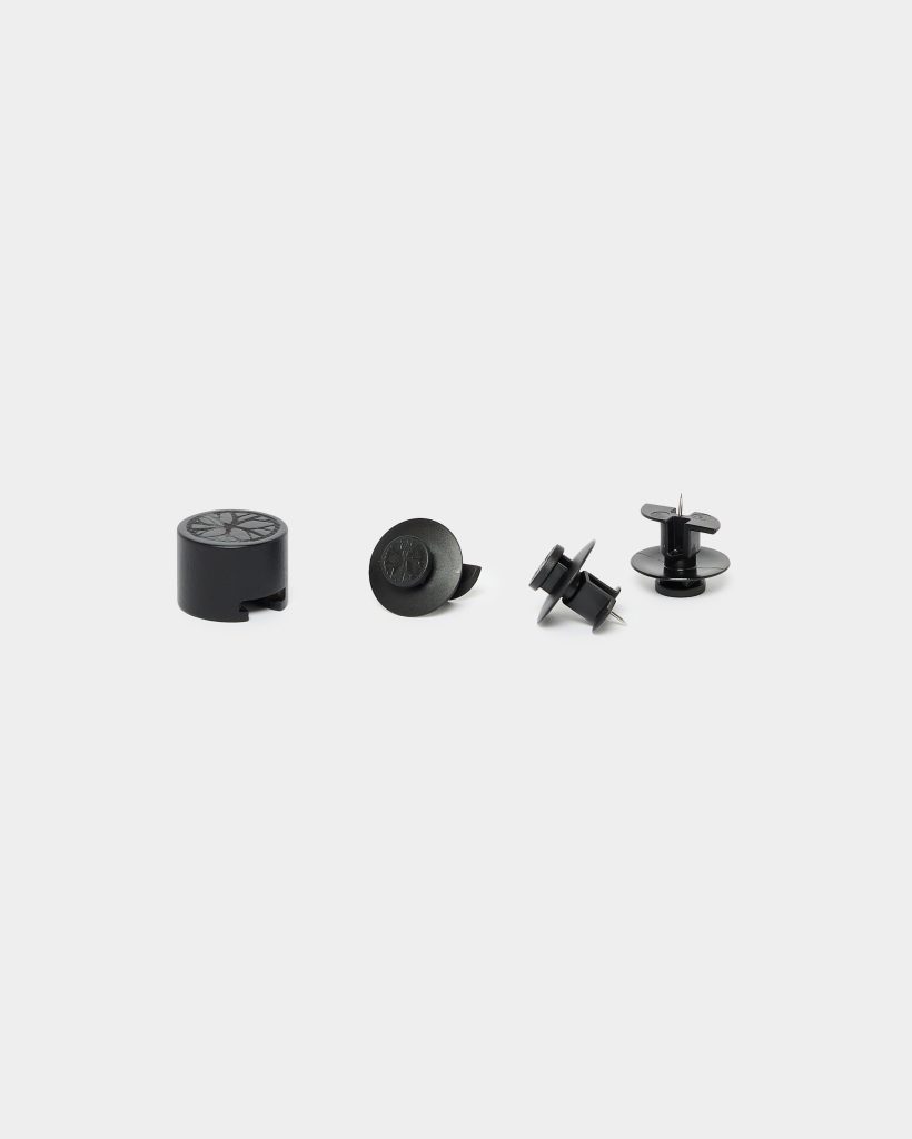 WoodUpp: four small black installation fittings arranged on a white background — a cylindrical plastic cap, a round snap-on cover, and two push-fit panel connectors with short metal pins. All items are matte black WoodUpp furniture fixings used for concealing screws and securing panels.