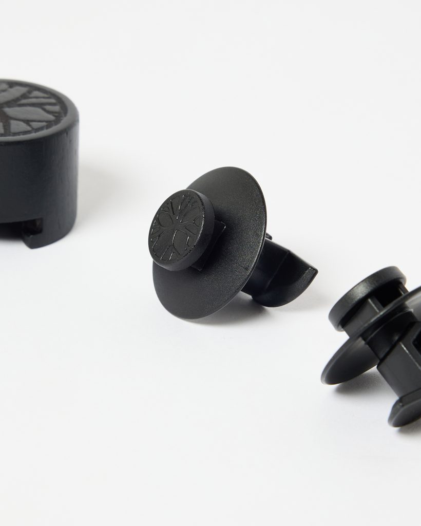 Close-up of WoodUpp products on a white background: two black circular car vent clips with disc-shaped fronts embossed with a stylized tree logo, plus a matching round wooden aroma puck. Minimalist plastic-and-wood design highlights a compact, matte black finish and ventilation mounting prongs.