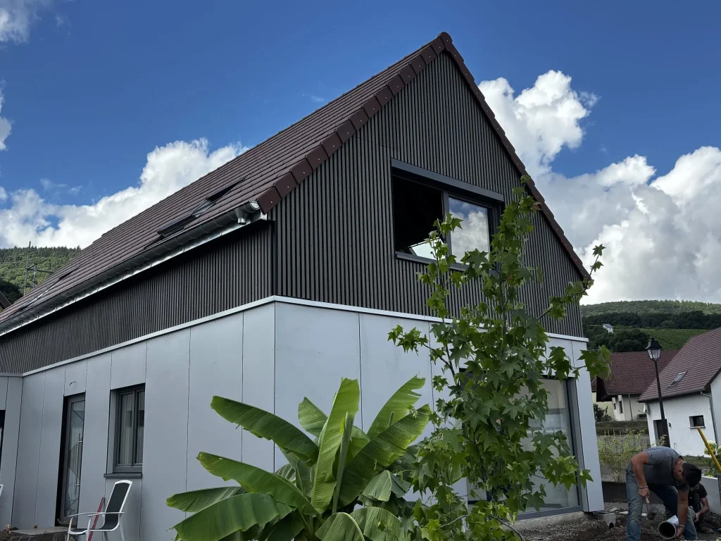 Exterior view of a modern two-story house featuring WoodUpp vertical dark timber slat cladding on the upper gable and WoodUpp smooth grey facade panels on the lower level, with a banana plant and young tree in front, a blue sky background and a worker at the right.