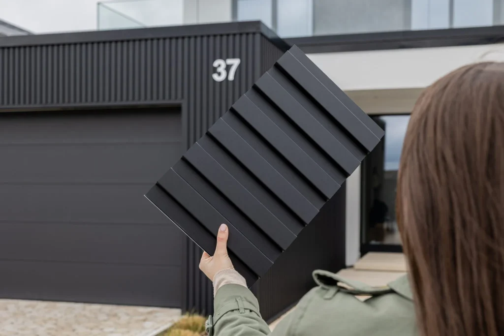 Person holds a WoodUpp black slatted cladding sample panel up against a modern house, showing the brand's dark ribbed exterior boards. The image highlights the textured vertical WoodUpp façade panels and matching dark garage door, illustrating color, profile and finish for exterior cladding selection.