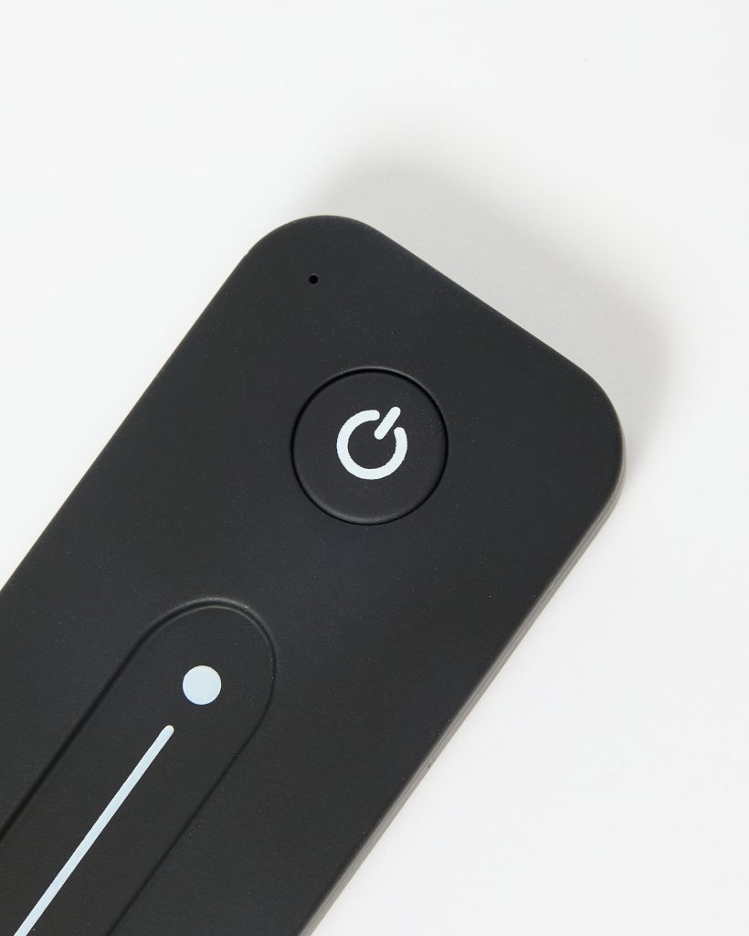 Close-up of a WoodUpp matte black wireless remote control showing a round power button with a white power icon and a long vertical touch-dimmer strip with circular position indicator; slim, rounded-corner rectangular design photographed against a white background and tiny status LED near the top edge.