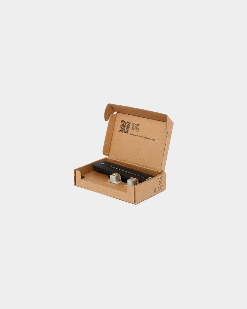 Open WoodUpp cardboard box at center of white background containing a black slim WoodUpp LED controller with power button and slider markings, plus two small metal mounting caps. Interior lid shows a QR code and printed setup icon; eco-friendly corrugated packaging holds components neatly.