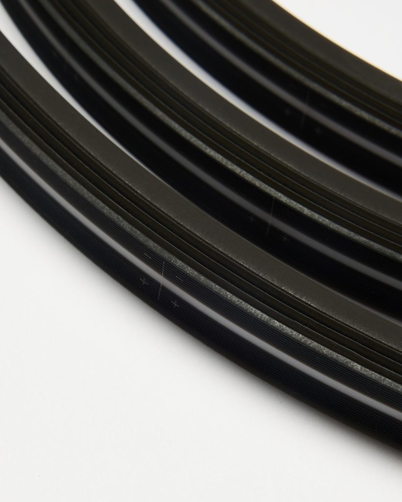 Close-up of WoodUpp coiled black edge trim: several flat, flexible plastic strips with fine parallel ridges, matte-to-gloss finish and a thin white accent stripe along the edge, shown layered in curved coils against a white background, highlighting texture and profile.