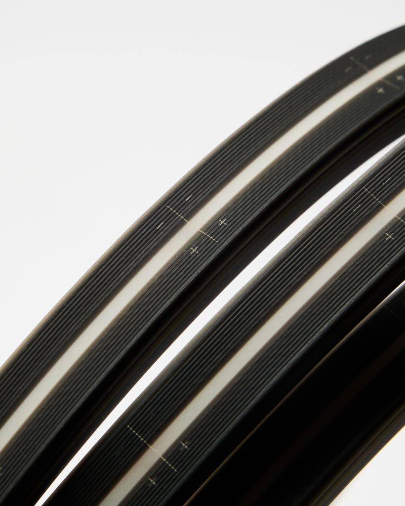 Close-up photograph of three curved WoodUpp wheel rim protectors: black ribbed rim strips with a central pale/white reflective stripe and printed alignment crosses. The WoodUpp products show textured grip ridges and smooth inner edges, presented against a bright white background.