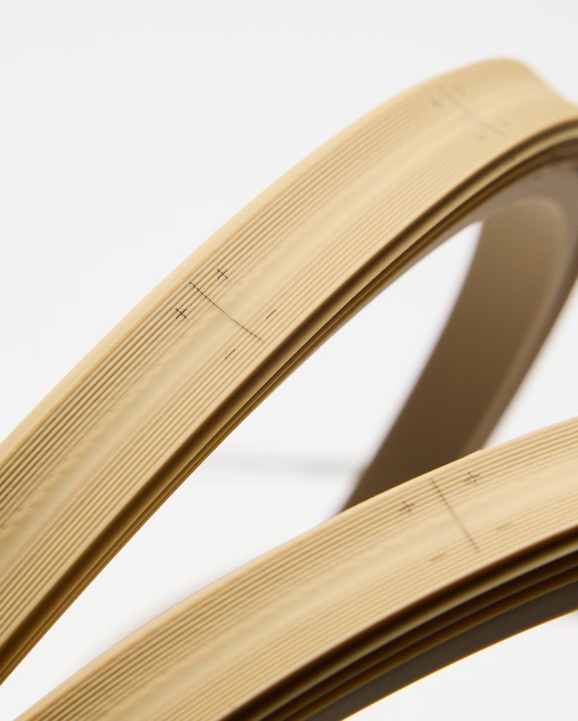 Close-up of two curved beige WoodUpp edge banding strips on a white background, showing fine parallel grooves, smooth adhesive-backed edges and printed alignment crosses and cut marks. The flexible, wood-tone strips are photographed in detail to highlight texture, trim guides and clean factory finish.