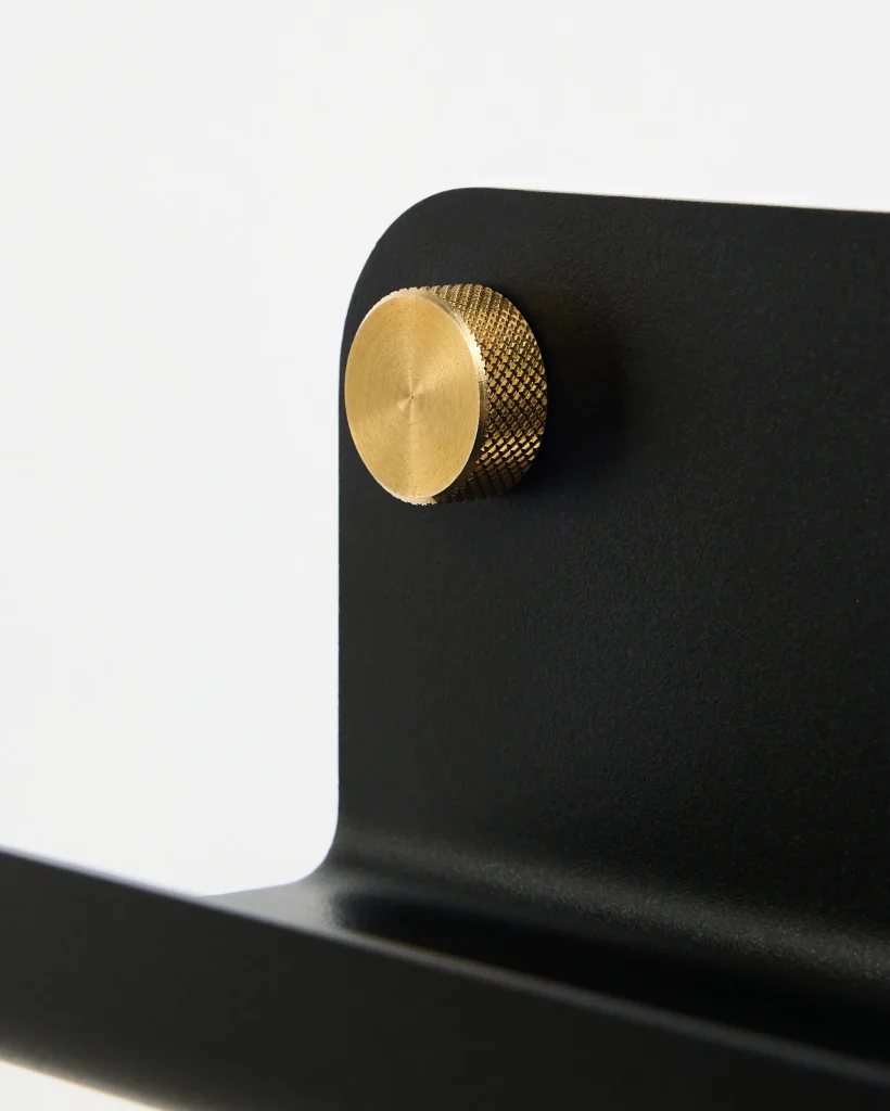 Close-up of a WoodUpp black metal wall hook featuring a matte, curved plate and integrated shelf lip, secured by a textured brass WoodUpp knob with circular brushed face. The image highlights the contrast between the black powder-coated steel and the knurled golden brass detail.