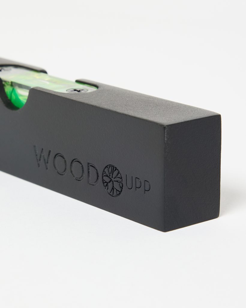 Close-up of a matte black WoodUpp compact spirit level, showing the embossed WOODUPP logo and circular tree-ring emblem on the wooden block, with a visible green bubble vial secured by black screws. The photo focuses on the level's chamfered corner against a white background.