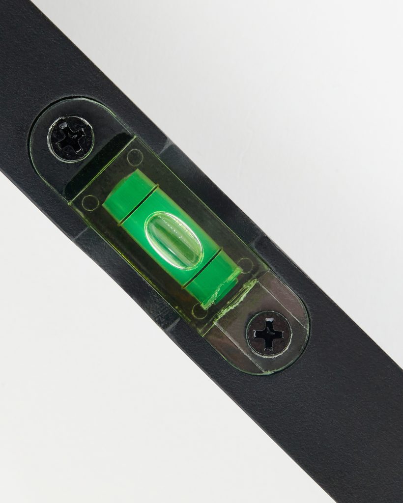 Close-up of a WoodUpp product showing a green spirit level vial integrated into a black metal bracket, the translucent capsule with a centered bubble, secured by two black Phillips screws. The compact WoodUpp bubble level insert aids accurate horizontal alignment on tools and fixtures.