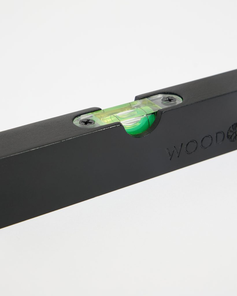 Close-up of a WoodUpp black wooden spirit level showing the translucent yellow-green bubble vial set into a rectangular black wooden frame, secured with two screws; the WoodUpp logo is embossed on the side, highlighting the brand's precision level tool design.