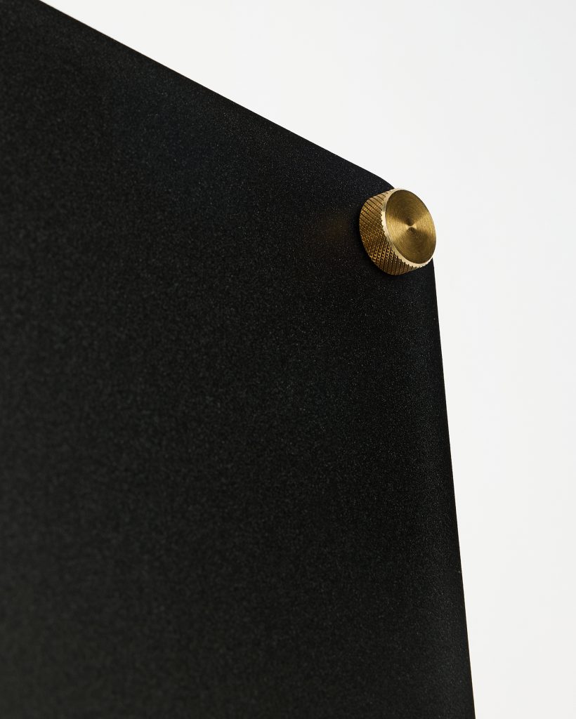 Close-up of a WoodUpp sleek black textured matte display panel, showing its fine-grain surface and angled corner, secured by a single brass knurled mounting screw—an elegant brass standoff with circular, textured edge used to fasten the panel to the wall.