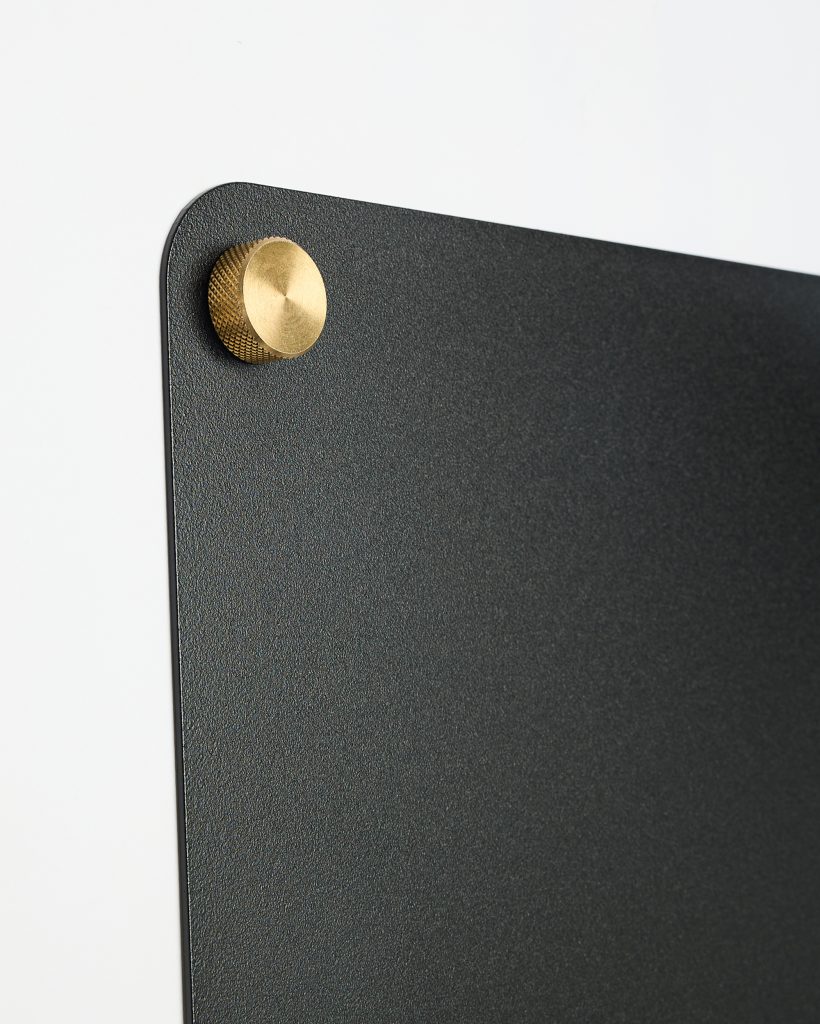 WoodUpp black textured wall panel with a rounded corner mounted on a white wall, secured by a single brass knurled standoff screw in the top-left. The image highlights WoodUpp’s minimalist matte black panel and decorative brass mounting knob with fine knurled detailing.