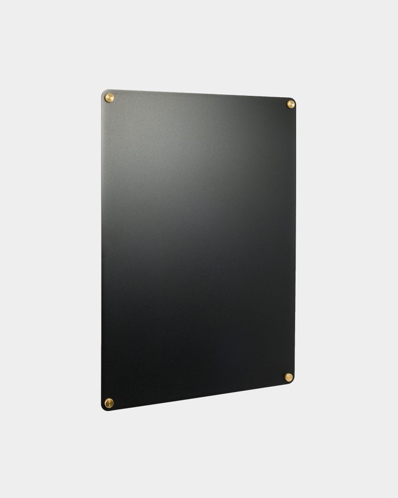 WoodUpp black rectangular wall panel with rounded corners photographed against a light gray background. The matte black board is mounted with four small brass circular standoffs, one at each corner, presenting a minimalist magnetic notice board from WoodUpp with an elegant brass finish.