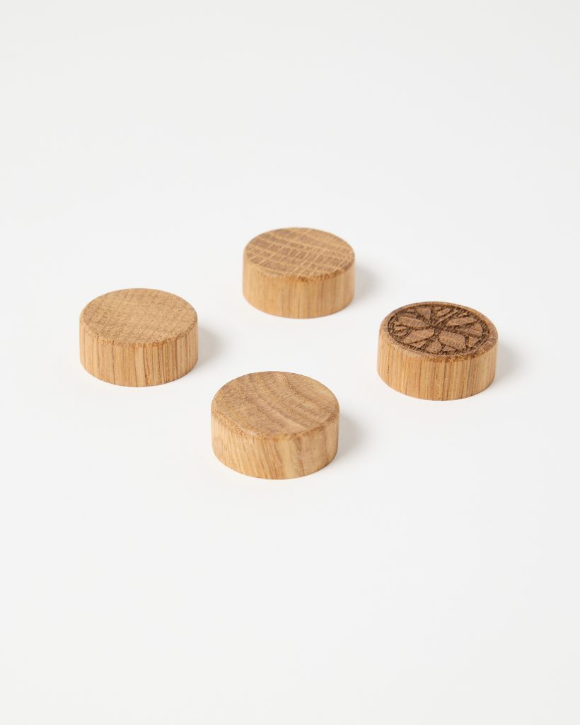 Four small round wooden plugs from WoodUpp are photographed on a white background: cylindrical oak screw-cover caps with smooth sanded sides and visible grain. Three are plain; one shows a stamped floral star pattern on top. They are natural light-brown, low-profile, and neatly finished.