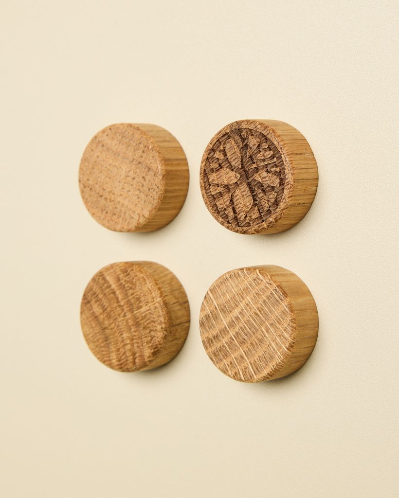 Four WoodUpp round oak magnetic wall hooks mounted on a pale beige surface, arranged in a neat square. Each knob displays natural oak grain and end-grain texture; one features a detailed laser-engraved tree motif. Smooth cylindrical profile and warm honey tones highlight the wood’s character.