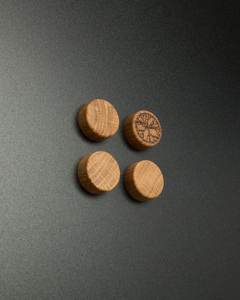 WoodUpp image showing four small round oak magnets on a dark textured surface: smooth cylindrical wooden plugs with visible grain, one engraved with a radial compass-like logo. The handcrafted WoodUpp wooden magnets emphasize natural oak texture, warm tones, and minimalist design.