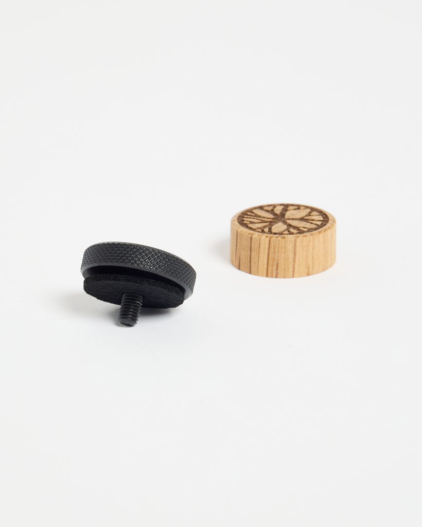 WoodUpp products: a black knurled metal thumb screw with felt pad and short threaded stud lies on a white surface beside a small round light-wood WoodUpp wooden cap featuring vertical grain and a laser-engraved circular floral pattern on top, shown in a minimal studio shot.