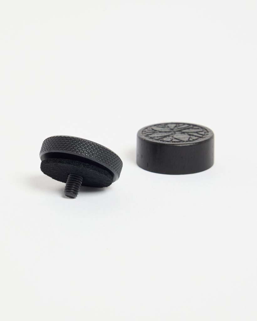 WoodUpp product image showing two black furniture accessories: a threaded adjustable glide with knurled metal edge, felt underside and short screw, and a solid round floor-protecting pad with a carved star-pattern top. Both items are matte black and photographed on a white background.