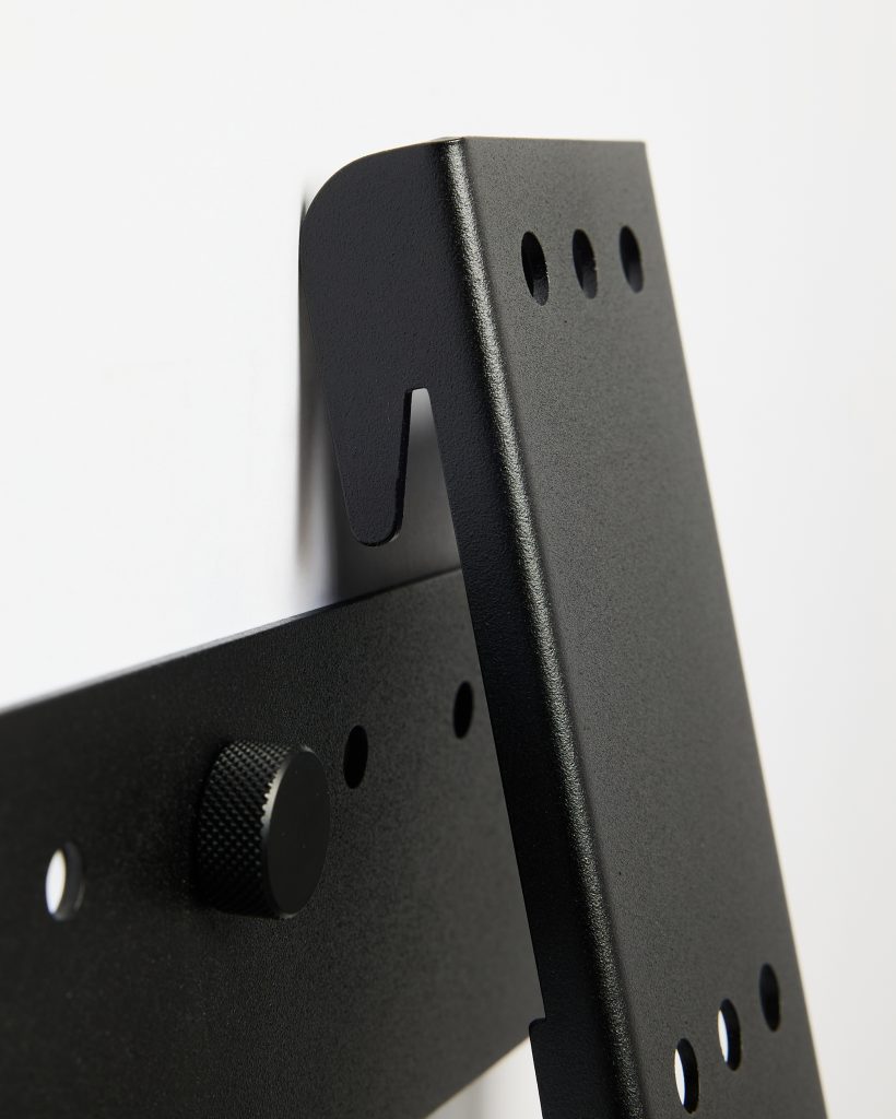 Close-up of WoodUpp black powder-coated metal wall-mount brackets: two interlocking flat plates with a hook slot, multiple VESA-style circular mounting holes, and a knurled adjustment knob, showing textured finish and precise cutouts designed for secure mounting of monitors and small TVs.