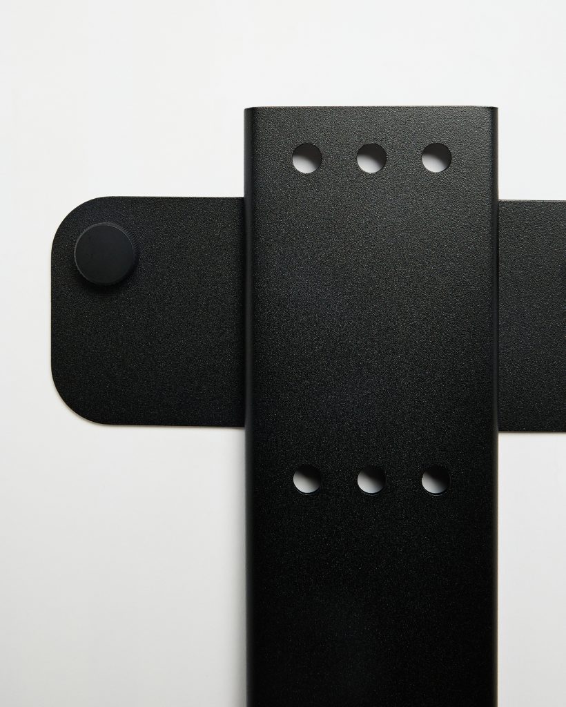 WoodUpp black metal mounting components photographed against a white background: a vertical, textured steel bracket with two rows of three circular holes and a horizontal rounded plate behind it featuring a knurled adjustment knob. Minimal, matte finish highlights the product’s clean, industrial design.