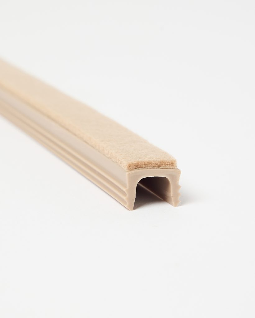 WoodUpp beige U-shaped door and window seal strip with adhesive felt pile: a flexible PVC rubber channel featuring grip ribs, a narrow tan felt top for soft contact, compressible interior profile for edge fitting and draught-proofing, shown close-up on a white background.