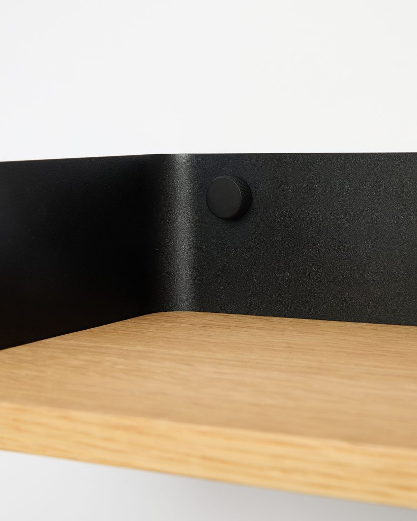 WoodUpp close-up of a light oak shelf with visible grain, mounted to a matte black powder-coated steel backplate; the curved bracket attaches with a textured round black screw cap. Minimal modern wall shelf detail highlighting WoodUpp’s oak shelf and black metal bracket components.
